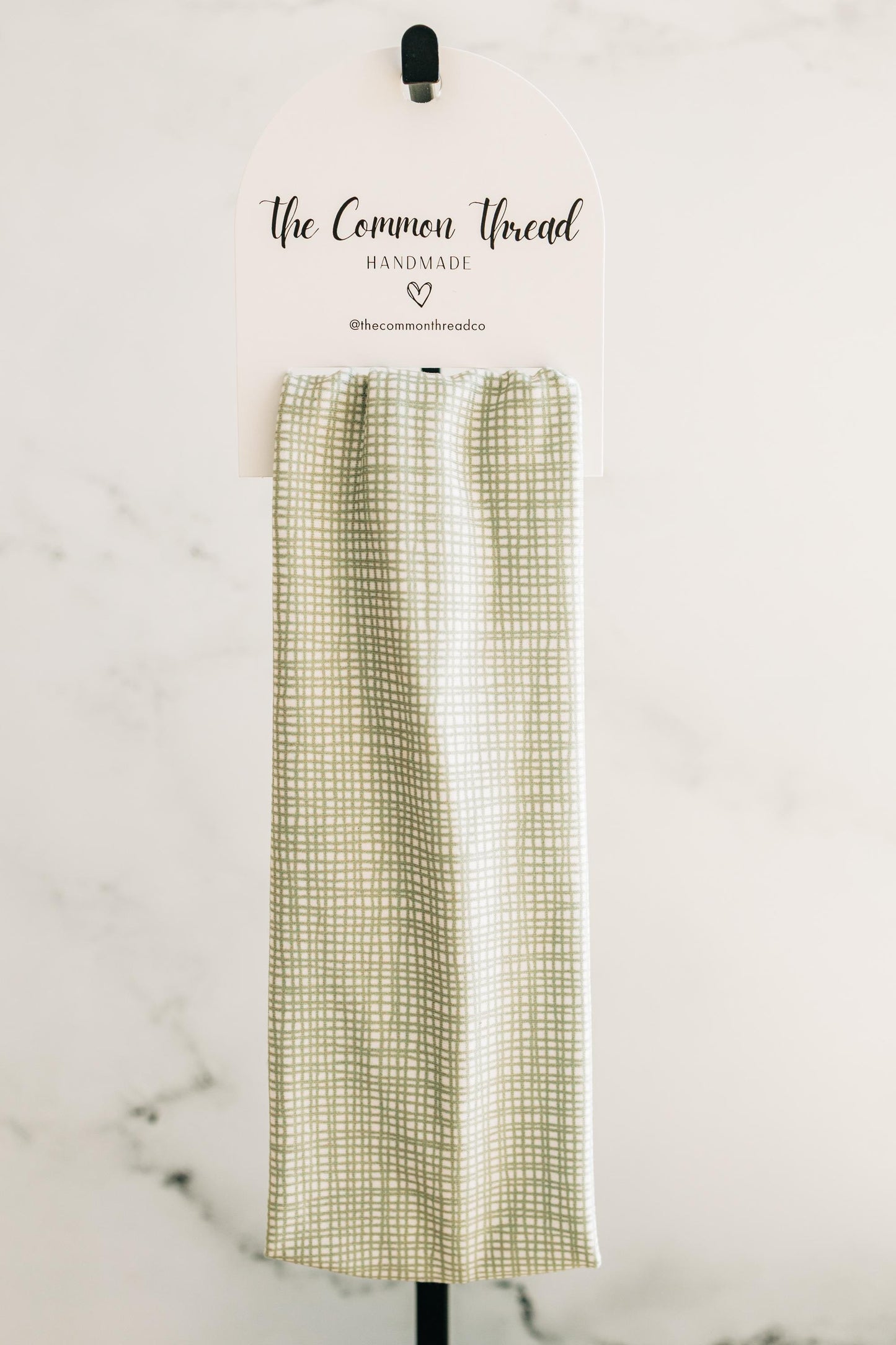May include: A light green and white checkered kitchen towel is displayed on a white sign with the text "The Common Thread Handmade". The towel is hanging from a black hook. The background is a white wall.