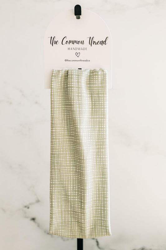 May include: A light green and white checkered kitchen towel is displayed on a white sign with the text "The Common Thread Handmade". The towel is hanging from a black hook. The background is a white wall.