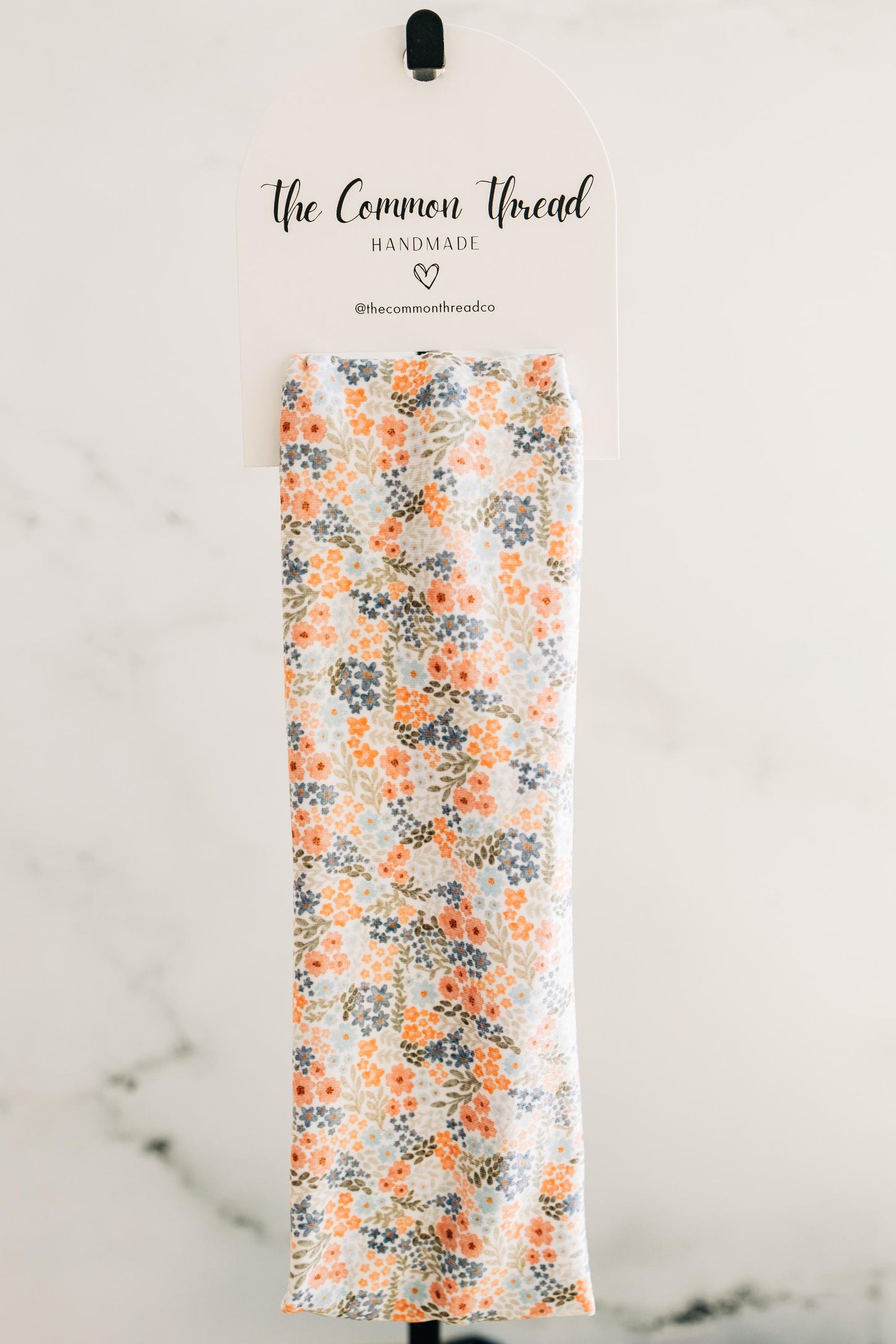 May include: A floral patterned fabric with orange, blue, and green flowers on a white background. The fabric is displayed on a white sign that reads "The Common Thread HANDMADE @thecommonthreadco".