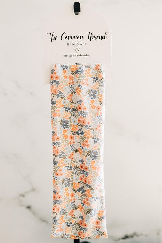 May include: A floral patterned fabric with orange, blue, and green flowers on a white background. The fabric is displayed on a white sign that reads "The Common Thread HANDMADE @thecommonthreadco".